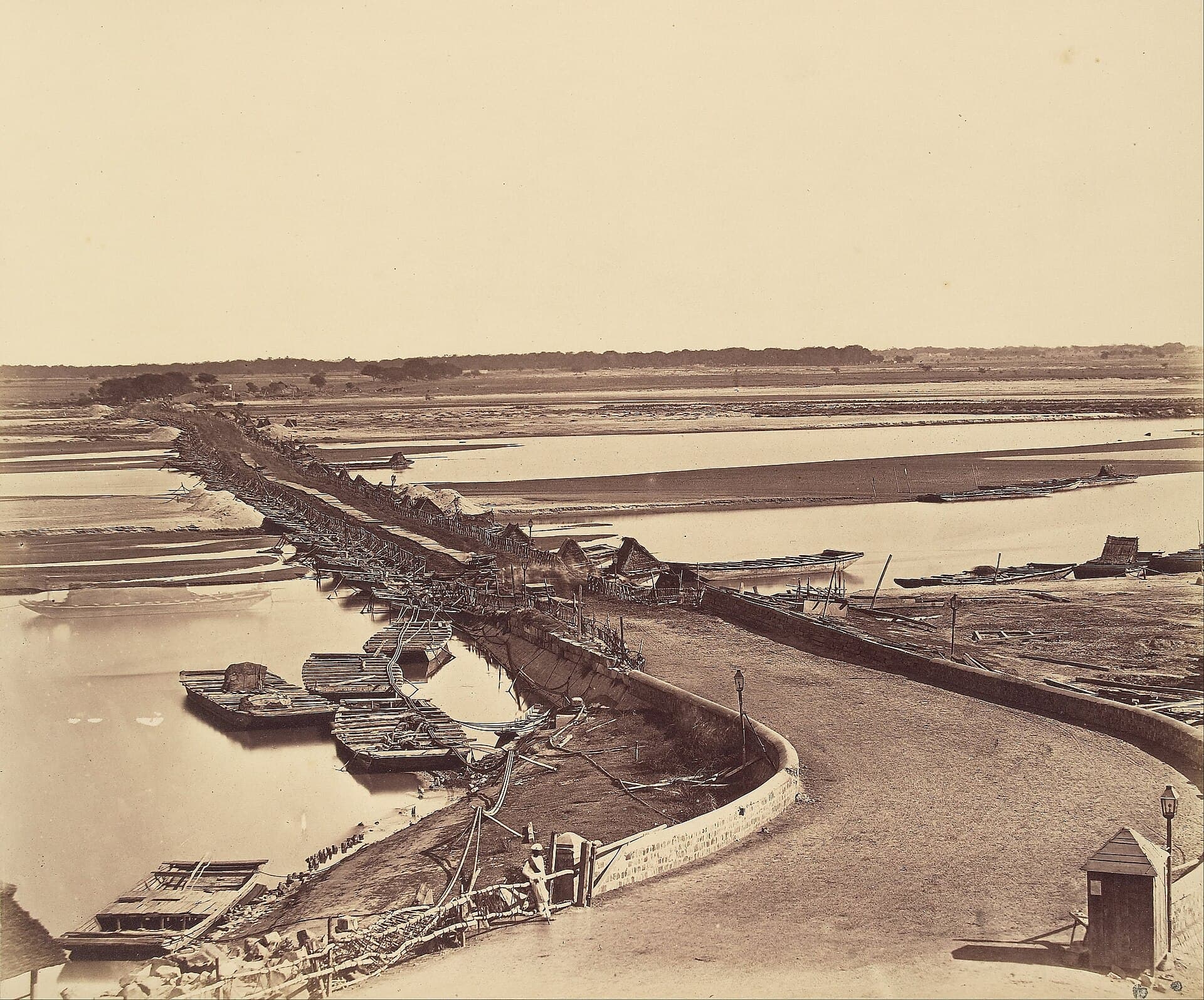 An 1858 albumen photograph of a bridge of boats crossing the wide, sandy Yamuna at Delhi, seen from the fort side of the river.