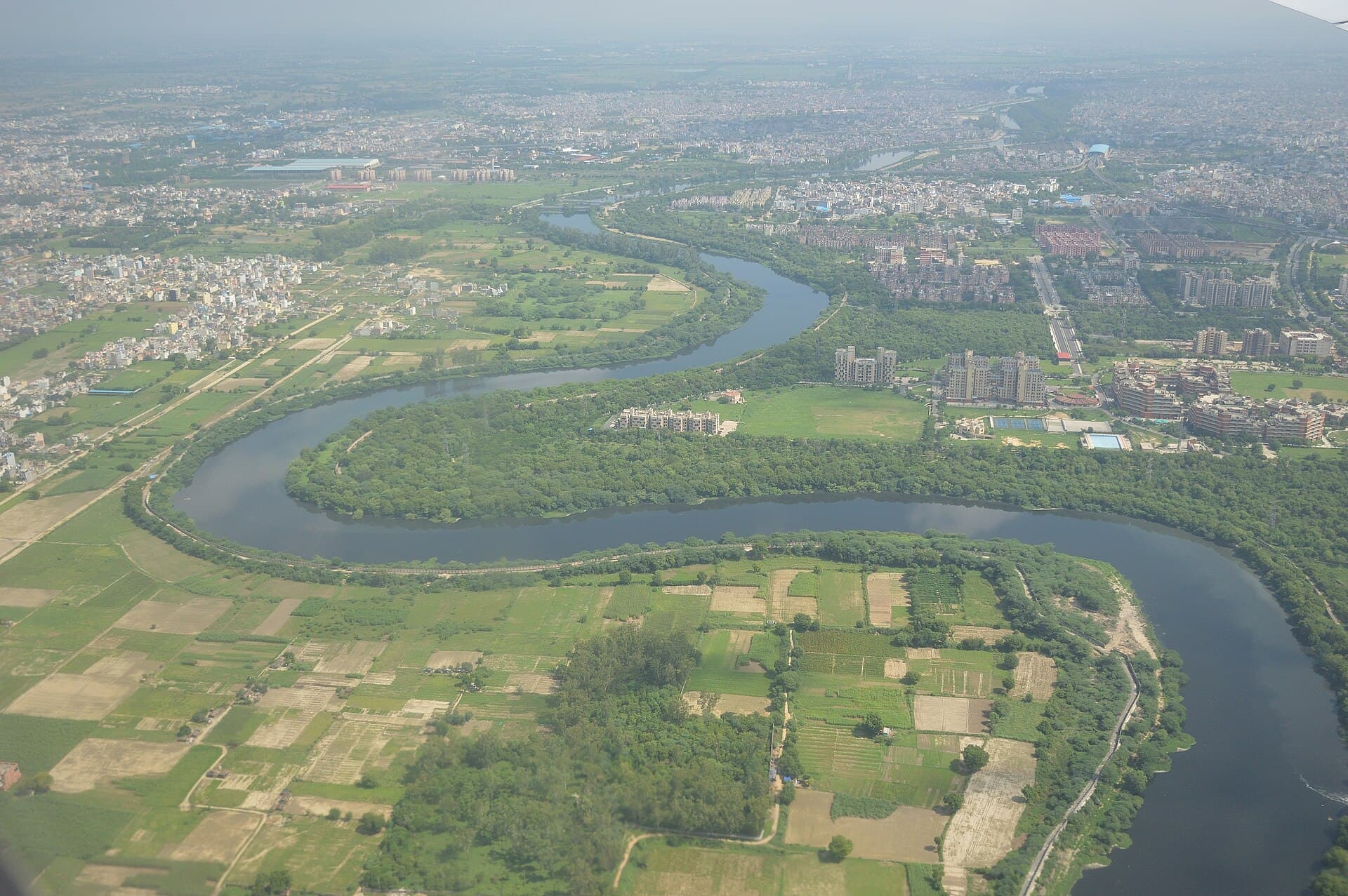 Aerial view of the broad, dark Najafgarh drain cutting through west Delhi's dense grey fabric of houses and roads.