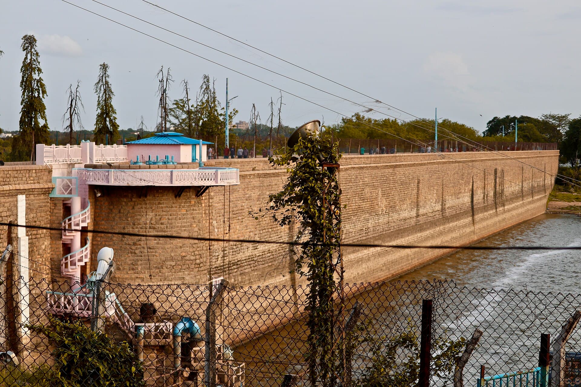 The masonry retaining wall of Osman Sagar reservoir at Gandipet, curving away from a pavilion at the near end, with the spillway channel and water to the right.