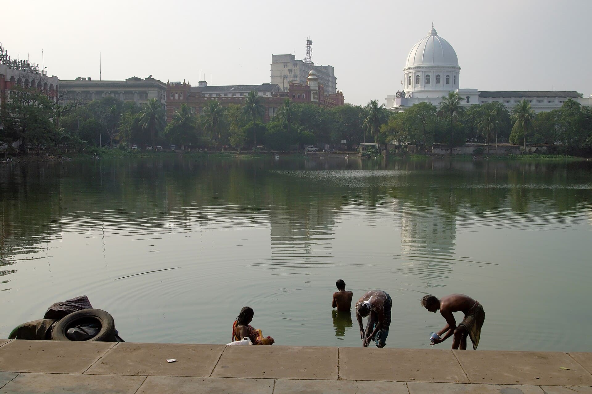 Lal Dighi in Kolkata: three people bathing and washing clothes at the tank's stone steps in the foreground, with the white dome of the General Post Office and the colonial administrative buildings of B.B.D. Bagh rising behind the water. A discarded tyre lies on the steps at the left.