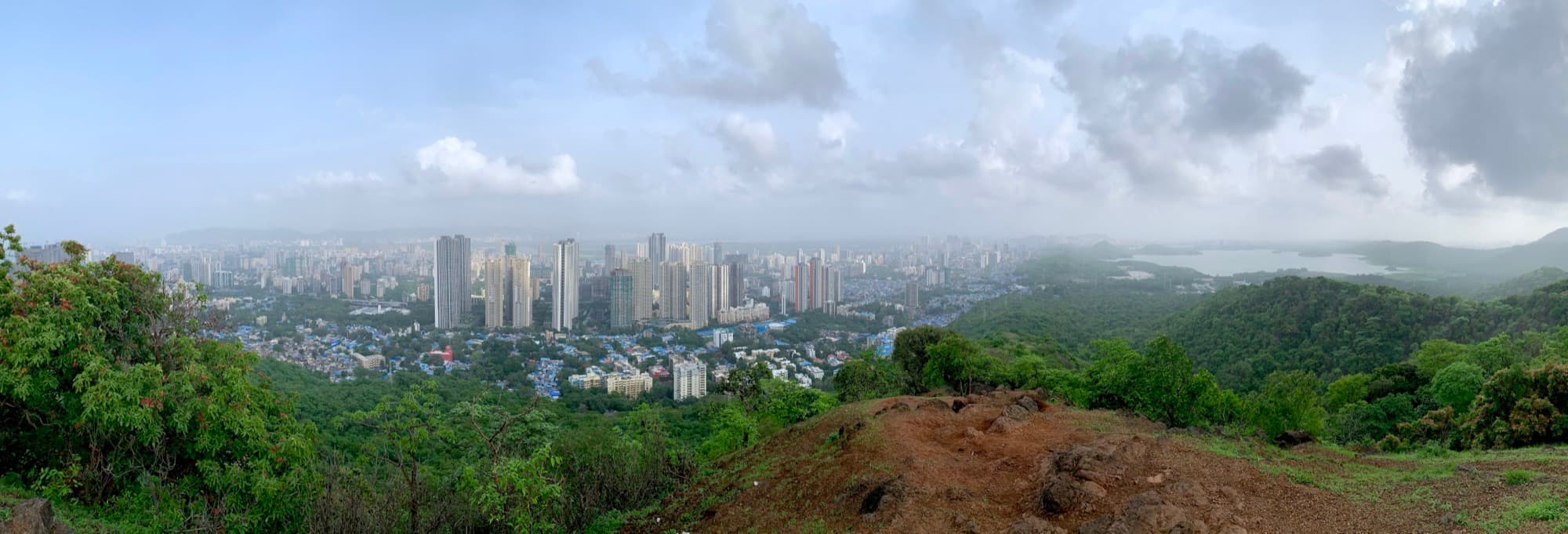 A wide panorama of Vihar Lake surrounded by forested hills, with the high-rise skyline of Mulund rising directly behind the ridge.