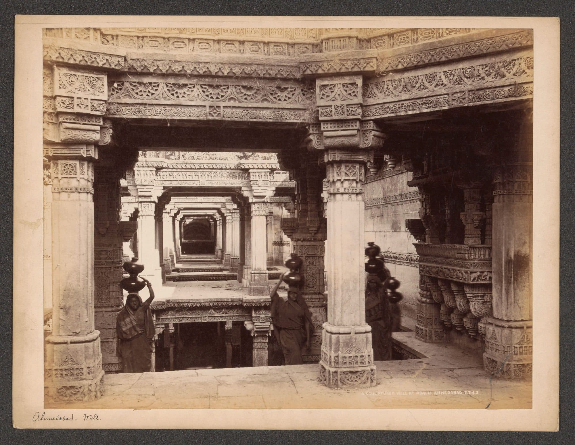 A sepia albumen print of the carved stone galleries of the Adalaj stepwell, with pillars and lintels framing a long view down the stepped corridor. Three women stand in the galleries carrying stacks of dark water pots on their heads. The print is captioned 'A Sculptured Well at Adalij, Ahmedabad. 2243'.