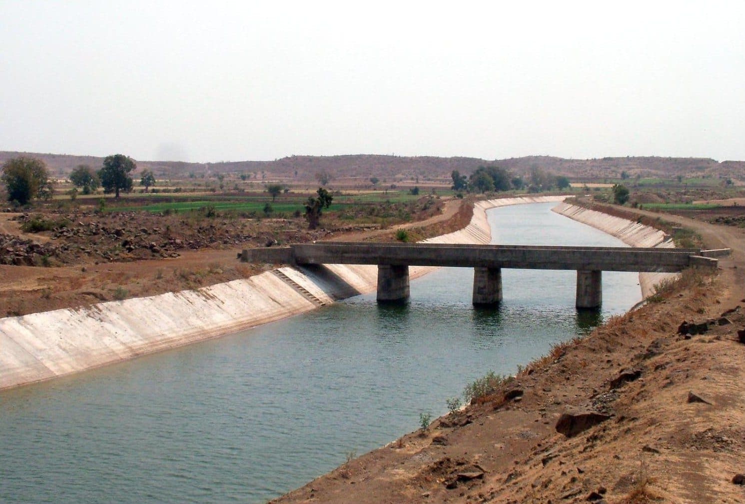 A wide concrete-lined canal running full of green water through dry, scrubby farmland, crossed by a low concrete bridge on three piers, with low hills on the horizon.