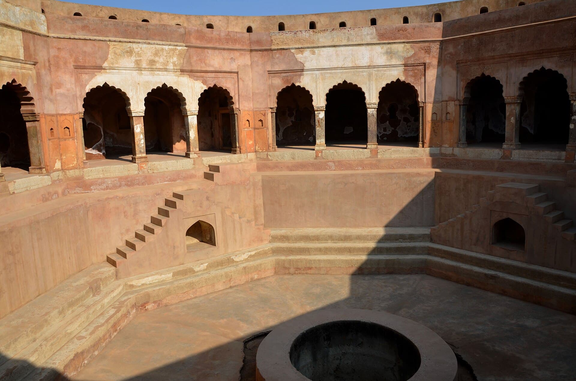 The interior of the Baoli Ghaus Ali Shah, an eighteenth-century stepwell at Farrukhnagar in Gurugram district. Curved sandstone galleries of cusped arches run around the upper level, flights of steps descend to a broad landing, and a circular well shaft opens in the floor at the bottom. The structure is completely dry.