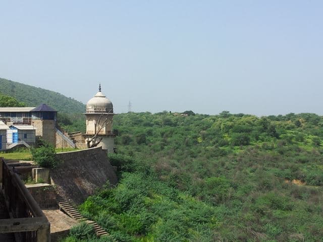 A view from beside a high masonry wall with steps and a small domed stone pavilion. Beyond the wall, low hills and a wide expanse of dense green scrub stretch to the horizon under a pale sky.