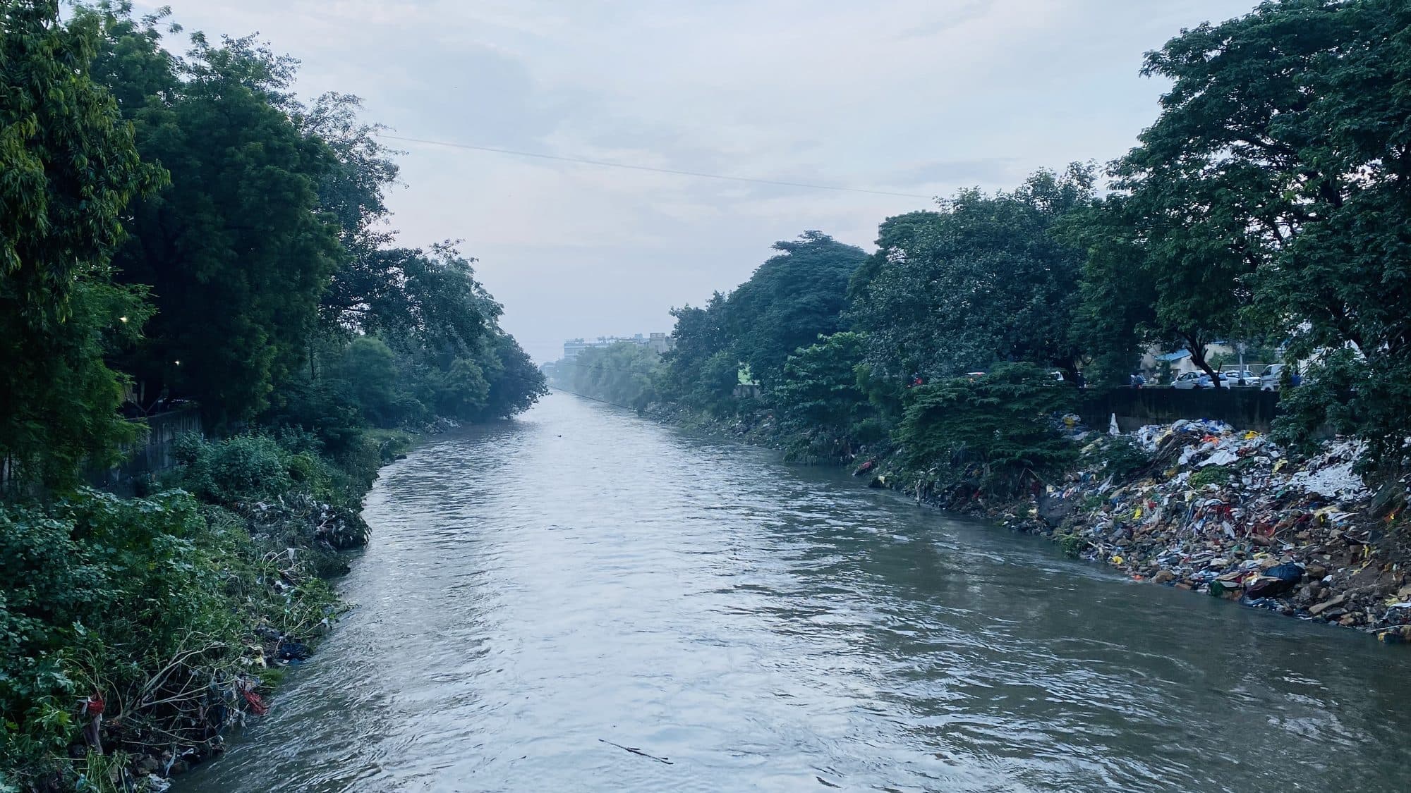 A fast, full brown river running straight between walls of trees. Rubbish and debris are piled high on the right bank below a wall, and a few cars are parked beyond it. Buildings stand in the distance under a grey sky.